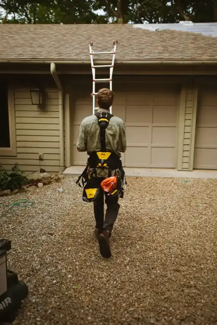 A roofer with a safety harness walking toward a ladder set against a house with a shingle roof
